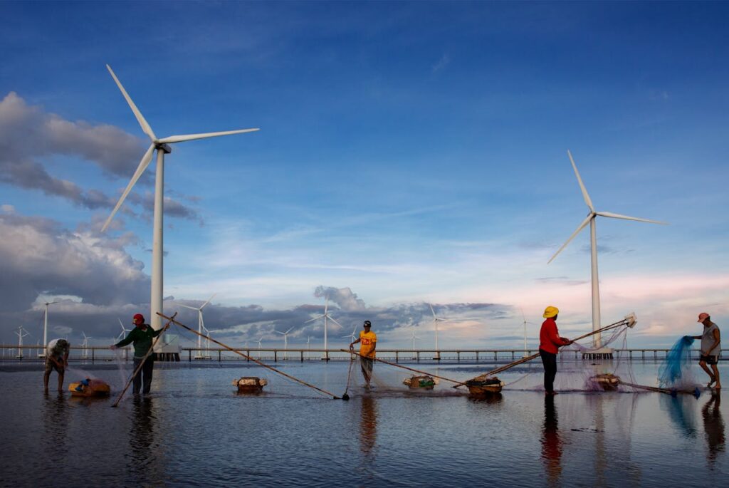 A group of fishermen cast nets near wind turbines set against a vibrant sunset sky, showcasing sustainable energy.