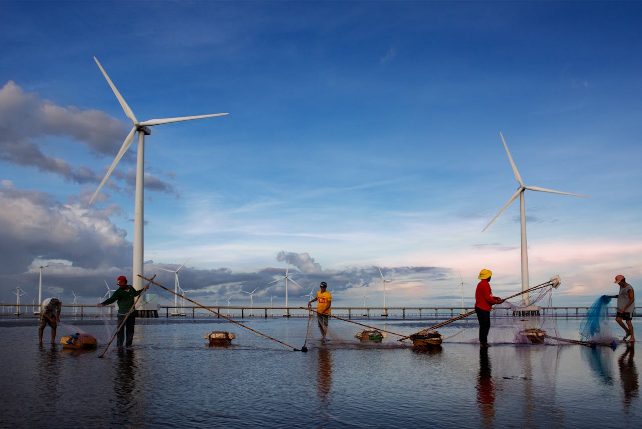 A group of fishermen cast nets near wind turbines set against a vibrant sunset sky, showcasing sustainable energy.